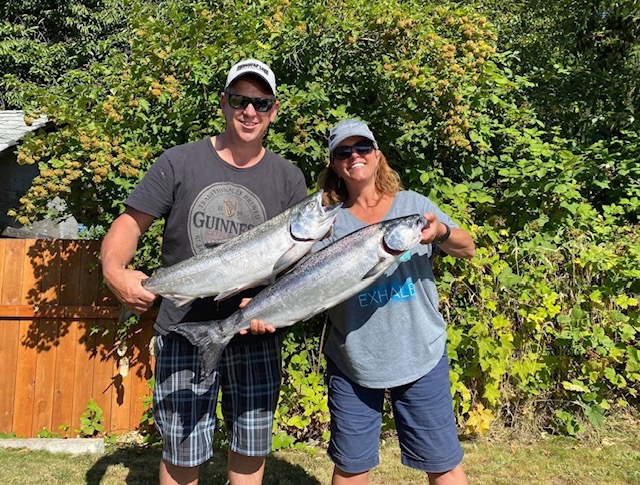 Two people holding a pair of fish in front of bushes and a fence. Both people are smiling, looking at the camera.